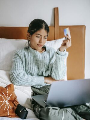 A young woman relaxes in bed, shopping online with a laptop and credit card in hand.
